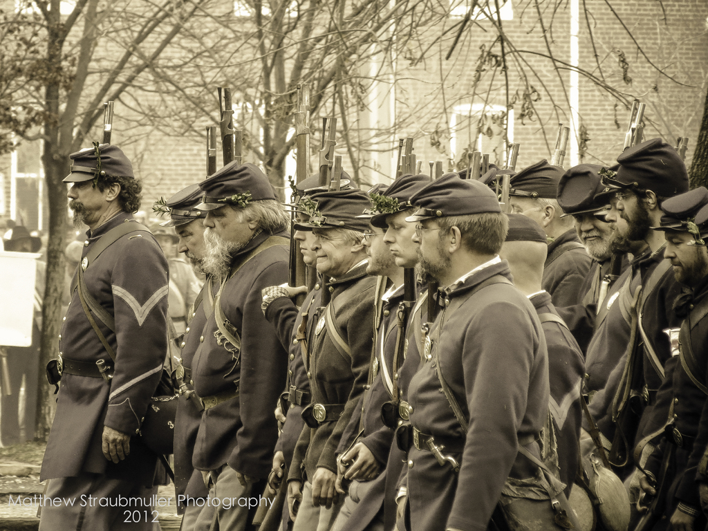 Mehrere Männer gehen auf einer Straße und tragen Mützen und halten Waffen; es gibt Bäume und ein Gebäude im Hintergrund; ein Wasserzeichen ist in der unteren linken Ecke sichtbar, schwarz-weißes Bild.