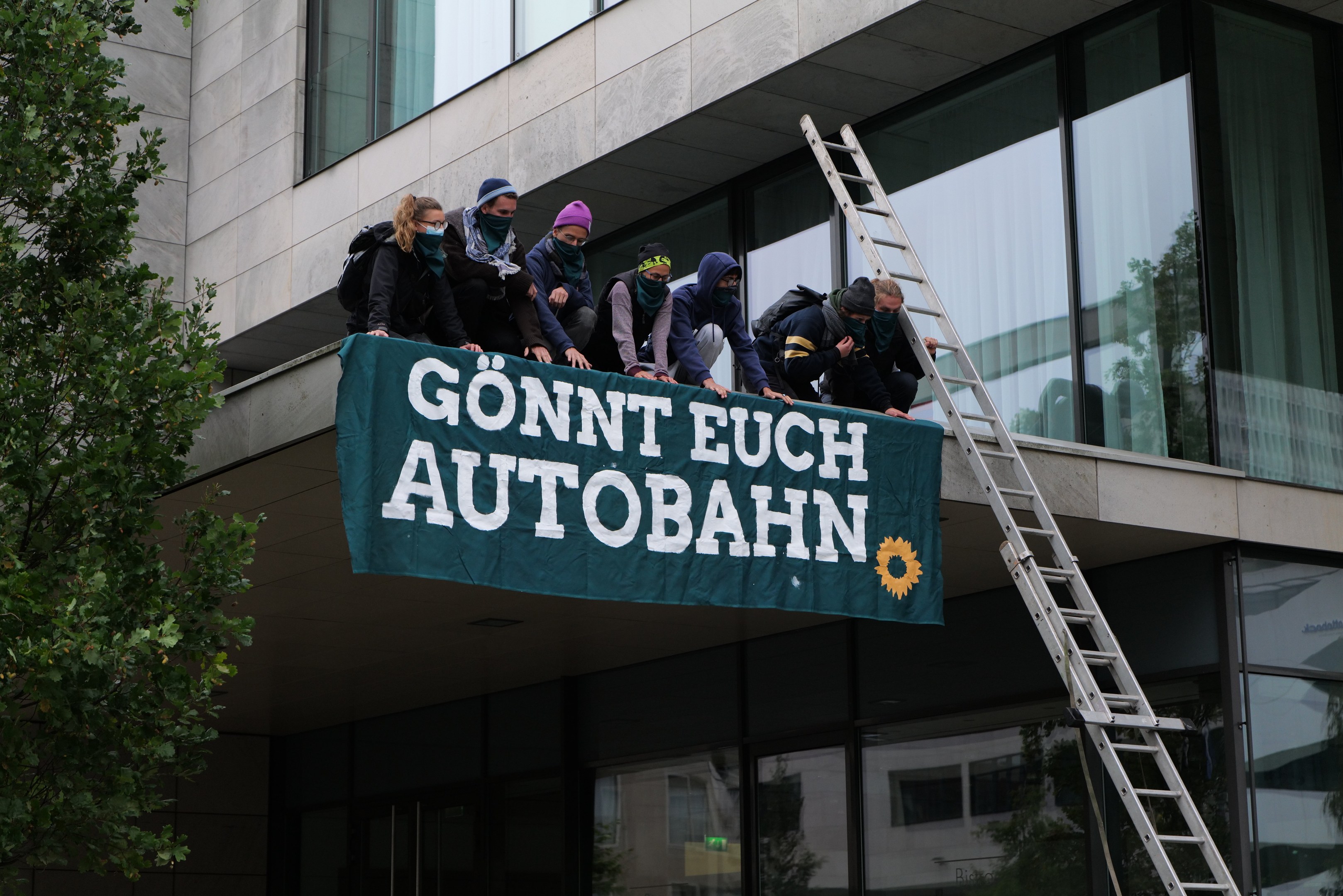 Eine Gruppe von Menschen auf einem Gebäude mit einer Fahne, mit einem Baum links und Glasfenstern im Hintergrund, die scheinbar für die deutsche Regierung protestieren.