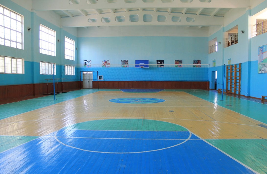 Großer Turnhalle mit blau-weißem Boden, Basketballfeld, Netz, Stangen, Tür, Fenstern, Wandplakaten, Geländer und Dach.