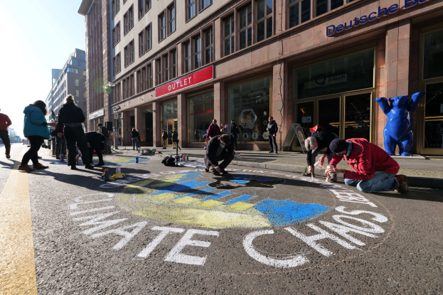 Menschen, die vor einem Gebäude auf dem Boden sitzen, während sie an einer Klimawandel-Demo in Berlin teilnehmen, umgeben von Flaschen und anderen Gegenständen, mit Bäumen und einem klaren blauen Himmel im Hintergrund.