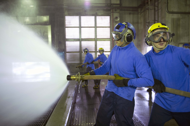 Eine Gruppe von Männern in Schutzausrüstung arbeitet an Maschinen in einer Fabrik, wobei einer Wasser auf den Boden sprüht.