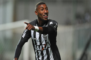 Ein Fußballspieler in einem schwarz-weißen gestreiften Trikot feiert mit einem Lächeln nach einem Torerfolg, der Hintergrund ist unscharf, um den Moment zu betonen.