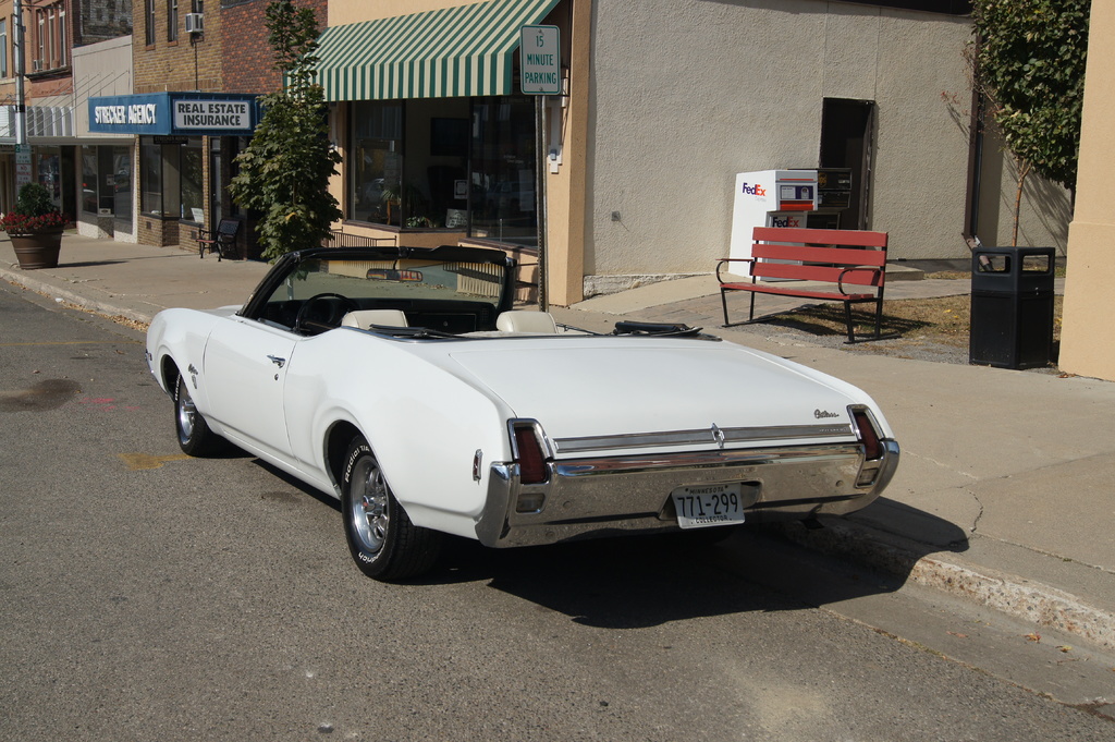 Ein weißes Auto mit der Nummer 771-299 steht auf der Straße, rechts daneben eine Bank, im Hintergrund ein Gebäude und ein 'Real Estate Insurance'-Geschäft.