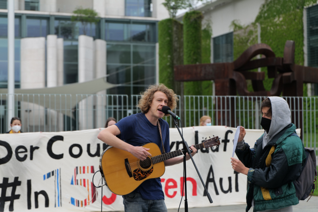 Ein Mann in einem blauen T-Shirt und Jeans spielt Gitarre und singt in ein Mikrofon auf einer Straße, während eine maskierte Person ein Blatt Papier zu seiner Rechten hält und ein Banner hinter ihm zu sehen ist, und eine kleine Menge sowie städtische Elemente wie Bäume und eine Skulptur im Hintergrund zu sehen sind.