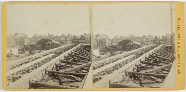 Schwarzes und weißes Foto einer Zugschiene mit Gebäuden im Hintergrund, Text 'Merkelbach & Co, Co. Amsterdam' rechts und hölzerne Gegenstände im Vordergrund verstreut.