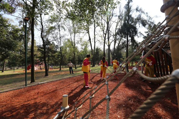 Gruppe von Menschen, die einen Seilparcours in einem Park mit Bäumen im Hintergrund und bodenbedecktem Gelände absolvieren, mit einer Stange mit Seilen auf der rechten Seite.
