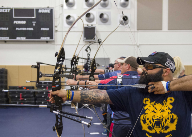 Eine Gruppe von Bogenschützen mit Mützen beim Hallentraining während der World Archery Championships 2017, mit einer Texttafel links und verstreuten Gegenständen rechts.