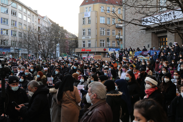 Eine große Gruppe von Menschen mit Mund-Nasen-Schutz steht vor einem Gebäude mit Fenstern, Bäumen, Schildern und Ampeln unter einem bewölkten Himmel, einige halten Schilder und Handys, scheinbar protestieren sie gegen eine Regierungsentscheidung, Masken zu verbieten.
