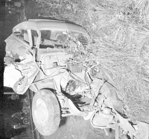 Schwarz-weiß-Foto eines schwer beschädigten Autos am Straßenrand mit eingedellter Front, zersplitterten Fenstern, einem Baum im Hintergrund und verstreuten Gegenständen.