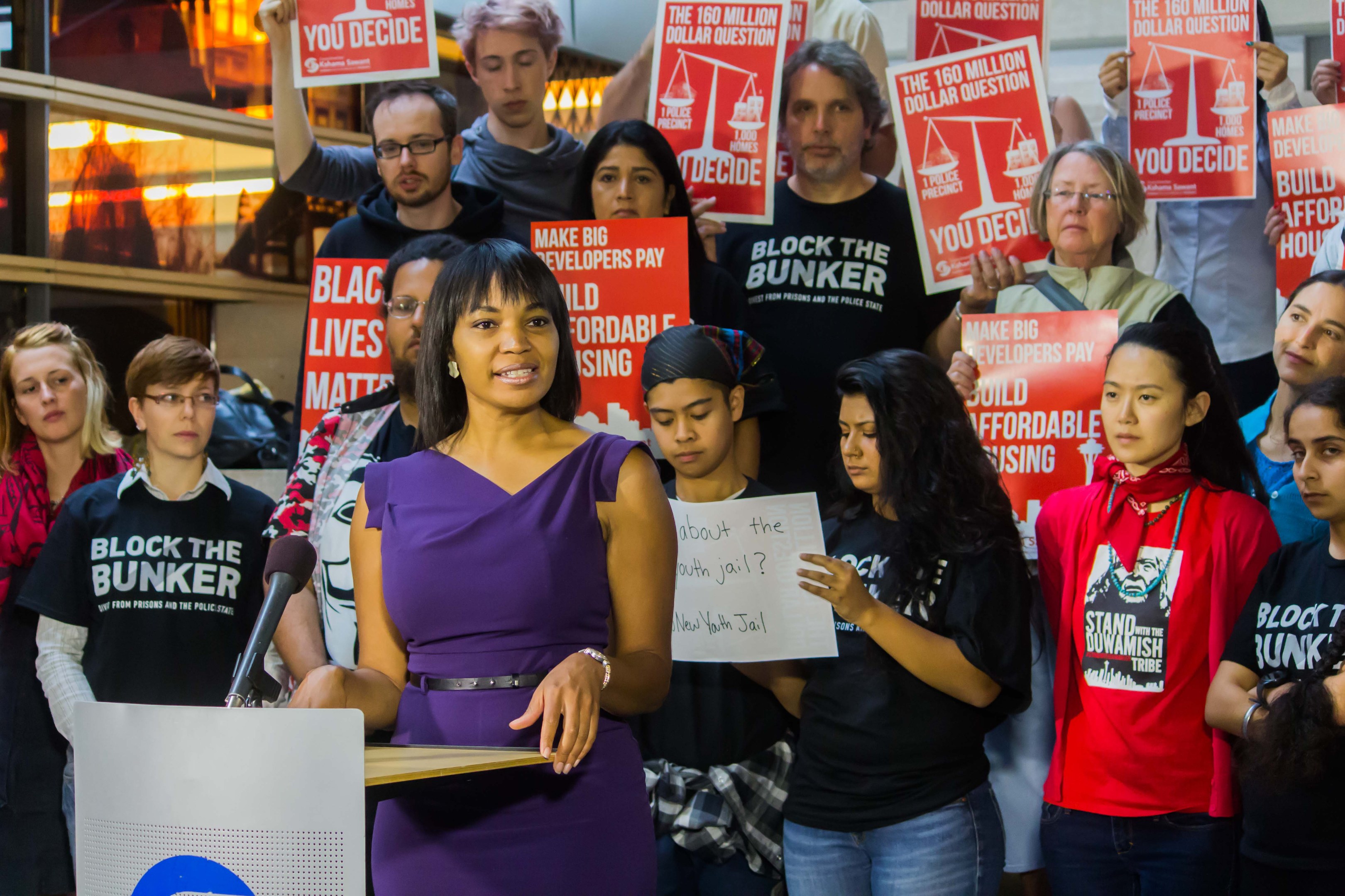 Eine Black-Lives-Matter-Aktivistin spricht an einem Rednerpult mit Mikrofon zu einer Menge, die Schilder hält, mit Lichtern und anderen Gegenständen im Hintergrund.