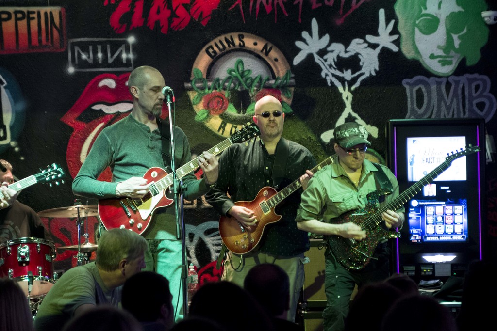 Drei Männer auf einer Bühne mit Gitarren, umgeben von einer Menge und einer bemalten oder gesprayten Wand im Hintergrund.