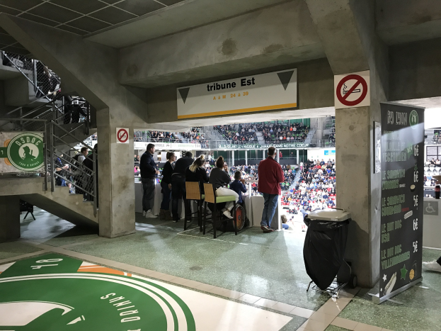 Eine Gruppe von Menschen steht vor einem sitzenden Publikum in einem Stadion, mit Schildern, die "Boston Celtics vs New York Jets" und andere Stadion-Elemente anzeigen.