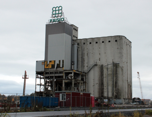 Großes Zementgebäude mit einem Zaun und umgeben von Vegetation, vor einem klaren blauen Himmel mit Bäumen im Hintergrund.