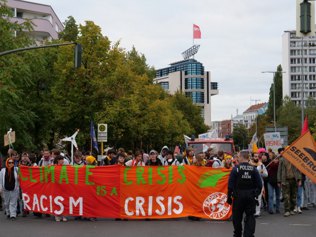 Eine Gruppe von Menschen mit einer "Klima-Krise ist eine Krise"-Plakette spaziert eine baumbestandene Straße mit geparkten Fahrzeugen, Schildern und Gebäuden unter einem klaren blauen Himmel entlang.