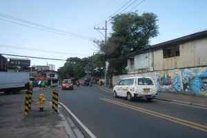 Eine städtische Straße mit fahrenden Fahrzeugen, einem Feuerhydranten, Pollern, Gebäuden mit Fenstern, Bäumen, Strommasten mit Drähten und einem Himmel als Hintergrund.