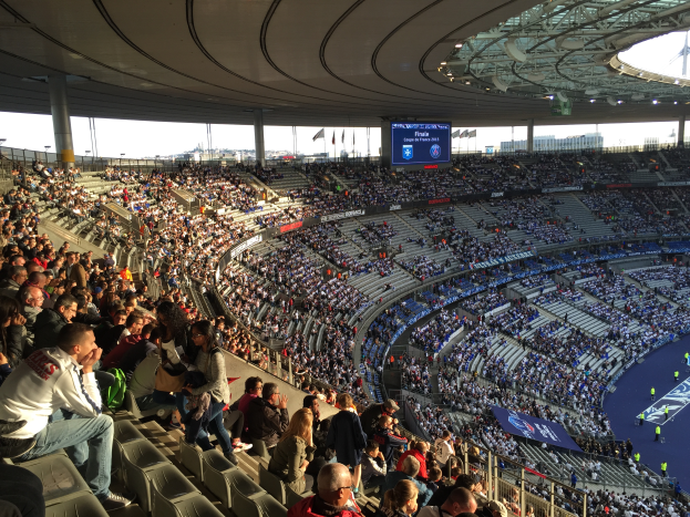 Eine große Menschenmenge sitzt im Allianz Stadion in München, Deutschland, und schaut ein Fussballspiel, mit einer Bühne auf der rechten Seite, Fahnen, Stangen und einem Bildschirm im Hintergrund und dem Himmel oben sichtbar.