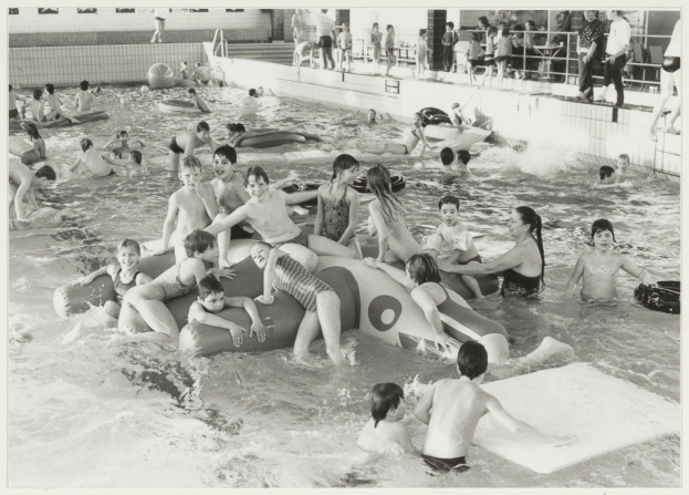 Schwarzes und weißes Foto von Kindern, die in einem Swimmingpool spielen, einige auf aufblasbaren Gegenständen und andere stehend, mit einer Wand und einem Geländer im Hintergrund.