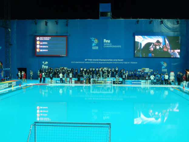 Eine Gruppe von Menschen, die um ein Hallen-Schwimmbecken mit Netz am Boden stehen, mit einer Wand im Hintergrund, auf der "FINA World Championships 2015 Kazan" angezeigt wird.