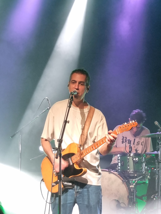 Ein Mann spielt eine Gitarre und singt in ein Mikrofon auf der Bühne, während ein anderer Mann Schlagzeug spielt.