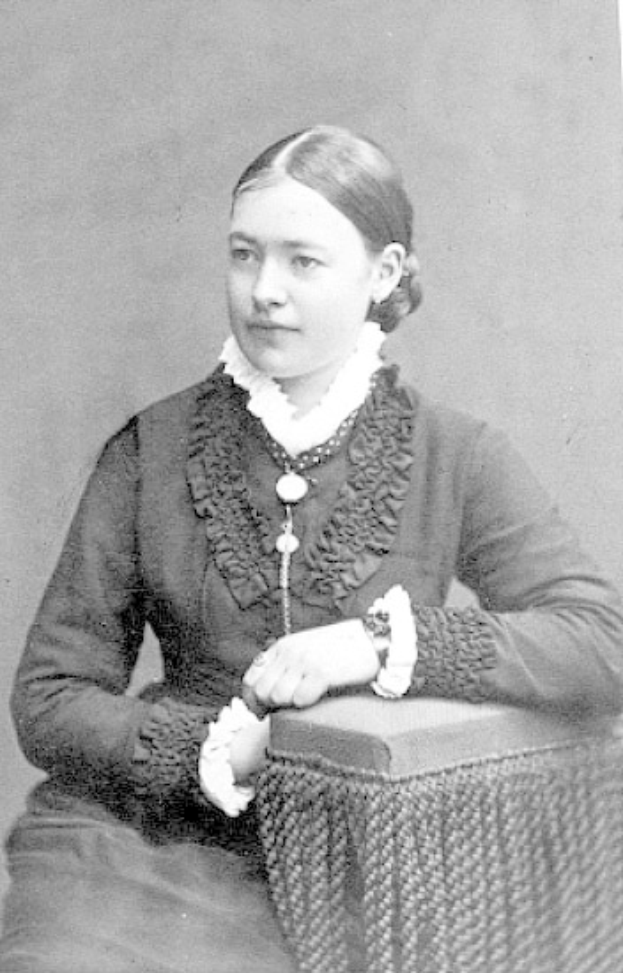 Schwarz-weißes Foto einer Frau auf einem Hocker sitzend, die ein Kleid trägt, mit zurückgekämmtem Haar und ernstem Gesichtsausdruck, die direkt in die Kamera schaut.