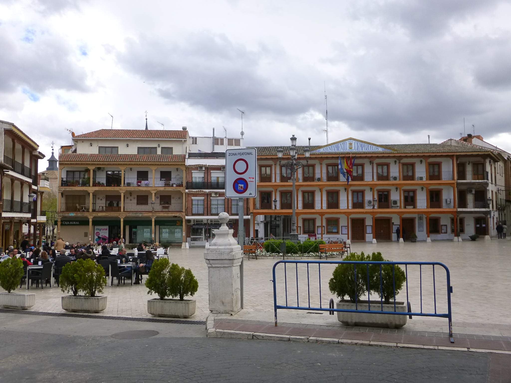 Ein belebter Stadtplatz mit Menschen auf Stühlen sitzend und stehend, umgeben von Topfpflanzen, Metallabsperrungen, Straßenlaternen mit Flaggen, einem Schild, Gebäuden mit Fenstern und einem bewölkten Himmel.