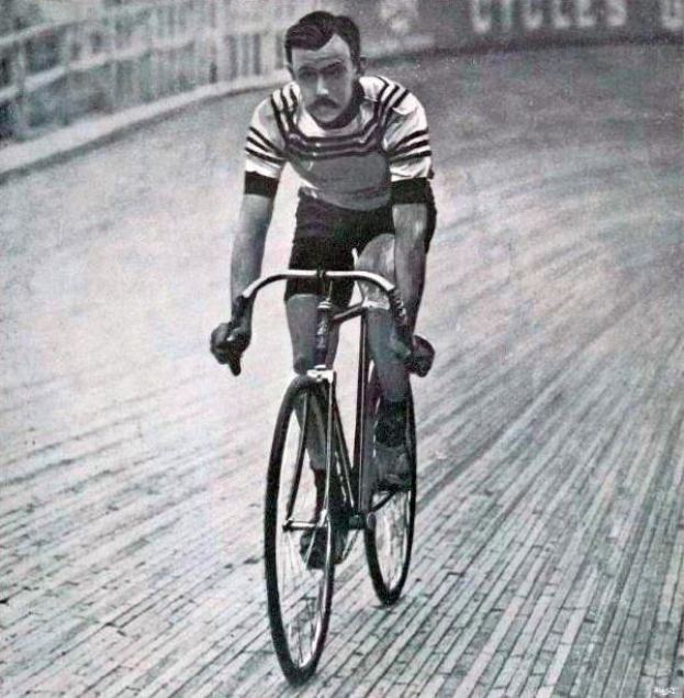 Altes Schwarz-Weiß-Foto eines Mannes, der auf einer Bahn mit einem Zaun und einer Tafel im Hintergrund Fahrrad fährt.
