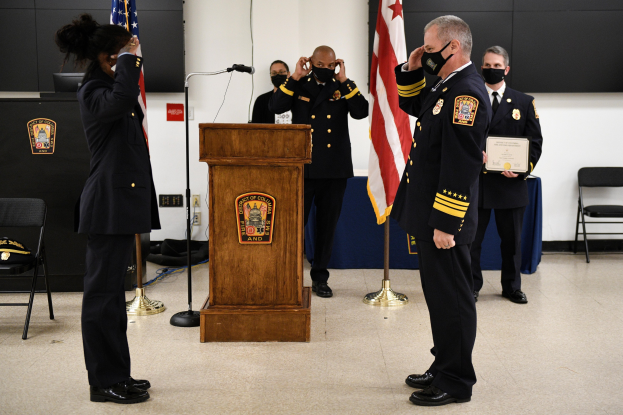 Gruppe uniformierter Personen in Masken vor einem Rednerpult bei einer Vereidigung eines neuen Feuerwehrchefs, der eine Urkunde hält, umgeben von Fahnen, Stühlen, Tischen und einer Wand.