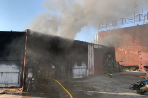 Feuerwehrleute in Helmen und Zylindern arbeiten daran, ein Feuer an einem Seecontainer zu löschen, während Rauch ausströmt und eine Person auf dem Boden sitzt und eine Pfeife hält und eine andere daneben steht, mit einem Gebäude, Fahnenmast und bewölktem Himmel im Hintergrund.