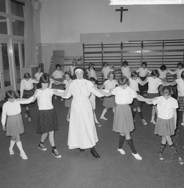Schwarzes und weißes Foto von Kindern in Schuluniformen, die in einem Raum mit einem Kreuzsymbol an der Wand, Glastüren mit Vorhängen und Deckenlampen tanzen.