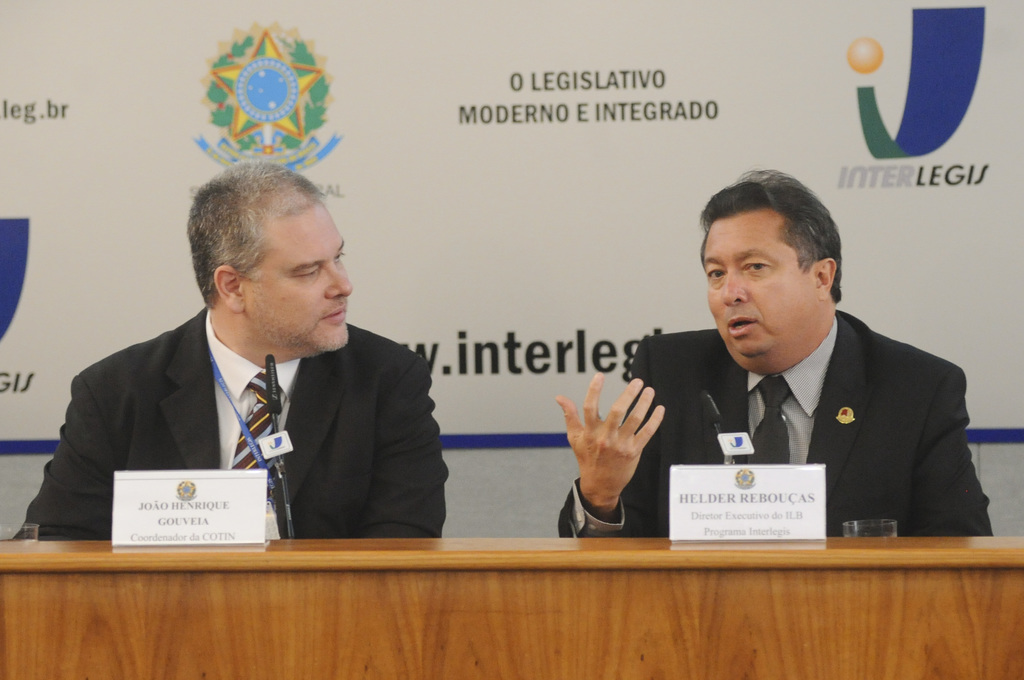 Zwei Männer in Anzügen sitzen an einem Tisch mit zwei Namensschildern und zwei Mikrofonen, mit Logos und Text im Hintergrund.