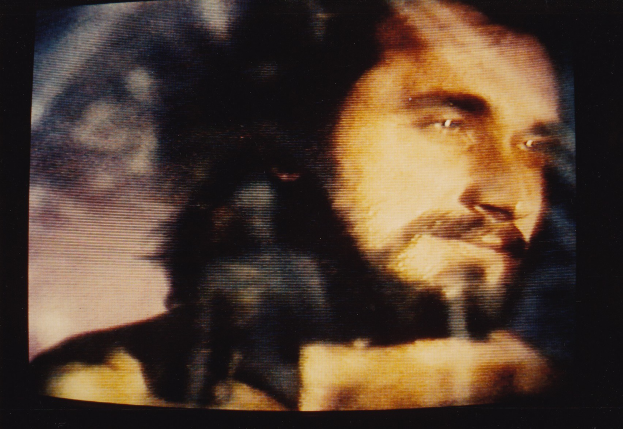 Ein Mann mit langem dunklem Bart und Schnurrbart, der ein weißes Hemd trägt, erscheint auf einem Fernseher mit dunklem Hintergrund und schaut direkt in die Kamera.