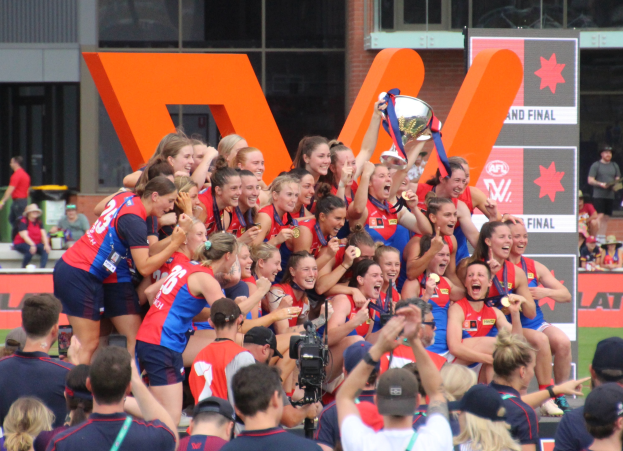 Gruppe von Frauen-AFL-Spielerinnen in blauen und roten Uniformen feiern mit einem Pokal vor einem jubelnden Publikum auf einem Rasenfeld.