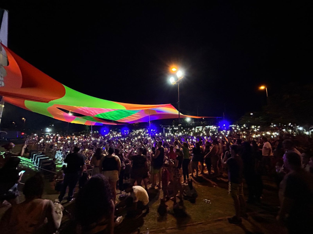 Eine große Menschenmenge steht vor einem Zelt in der Nacht, beleuchtet von bunten Lichtern und umgeben von beleuchteten Pfählen, mit einem Gebäude links und dem Nachthimmel im Hintergrund.