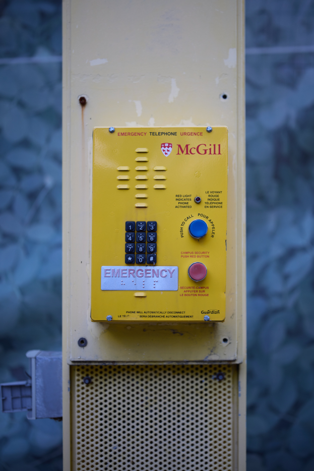 Ein gelber Notrufhörer an einer Gebäudeseite angebracht, mit Knöpfen und Text zum Absetzen von Notrufen, vor einer Wand.