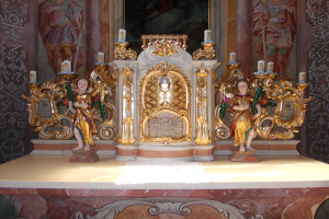 Ein Altar in einer Kirche mit Skulpturen von Menschen und Kerzen auf Ständern, vor einer Wand mit einem Gemälde, schafft eine feierliche Atmosphäre.