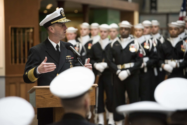 Ein Mann in militärischer Uniform spricht an einem Podium während einer Zeremonie mit Menschen in weißen Mützen, während bewaffnete Personen hinter ihm stehen, in der Nähe einer Fahne und Lichter.