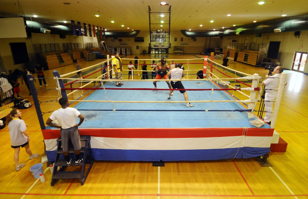 Gruppe von Menschen um einen Boxring in einer Turnhalle mit einem Fotografen auf der rechten Seite und Deckenlampen, die eine professionelle Atmosphäre suggerieren.