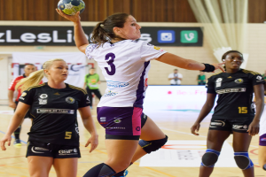 Eine Gruppe von Frauen spielt Hallenhandball, wobei eine den Ball hält, in einer Halle mit Zuschauern und Werbetafeln im Hintergrund.