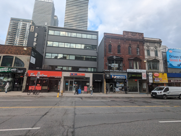 Stadtstraße mit hohen Gebäuden, fahrenden Fahrzeugen, Fußgängern, einem Fahrrad, einem Feuerhydranten und Werbetafeln für Mietwohnungen unter einem bewölkten Himmel.