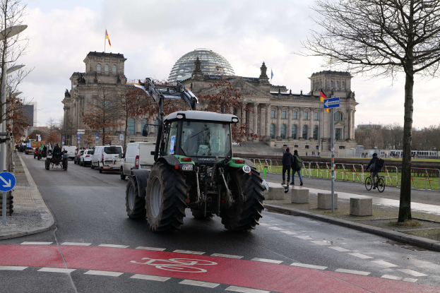 Ein Traktor fährt eine Straße mit Bäumen, Laternen und Schildern entlang und passiert das Reichstagsgebäude in Berlin, Deutschland, das mit Flaggen geschmückt ist, während Menschen und Fahrradfahrer auf dem Gehweg unter einem bewölkten Himmel unterwegs sind.