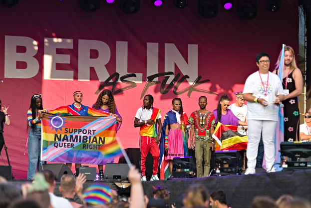 Eine Gruppe von Menschen auf einer Bühne mit einem Banner, auf dem "Berlin as Fuck" steht, mit Lautsprechern, Mikrofonen und anderen Gegenständen drumherum und einer feiernden Menge darunter, bei der einige Hände in die Höhe gestreckt werden.
