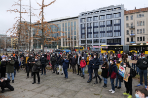 Eine große Gruppe von Menschen, einige mit Masken und Schildern, steht vor einem Gebäude während einer Protestaktion in Berlin. Es sind Fahrzeuge, Bäume, Laternenpfähle und Gebäude mit Fenstern im Hintergrund zu sehen sowie ein sichtbarer Himmel. Einige Personen halten Kameras.
