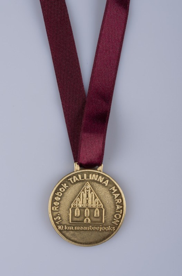 Goldmedaille mit einer maronenfarbenen Schleife, die mit "Reebok Tallinn Marathon" beschriftet ist, vor einem weißen Hintergrund.