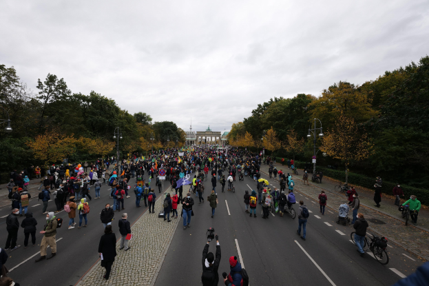 Eine große Gruppe von Menschen marschiert eine von Bäumen gesäumte Straße in Berlin entlang und hält Kameras in der Hand, mit einem Gebäude und einem klaren Himmel im Hintergrund.