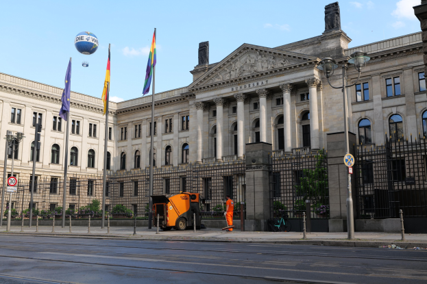 Großes modernes Gebäude mit zahlreichen Fenstern, als Bundestag in Berlin, Deutschland identifiziert, umgeben von Straßeninfrastruktur, Fußgängern und Flaggen, vor einem bewölkten Himmel.