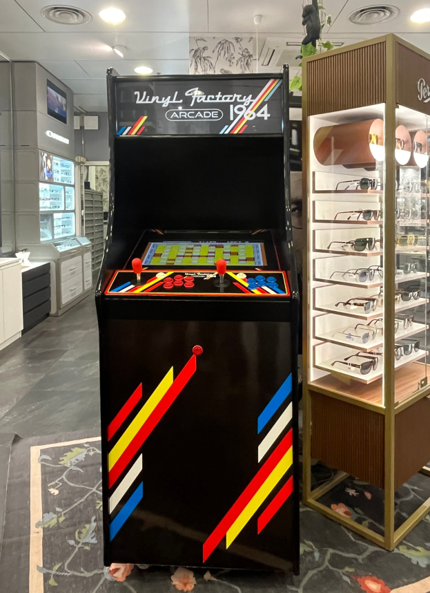 Ein Geschäft mit einem zentralen Retro-Arcade-Automaten, einem Glastisch auf der rechten Seite und einer retro-stylischen Beleuchtung mit Schränken und einem Fernseher im Hintergrund.