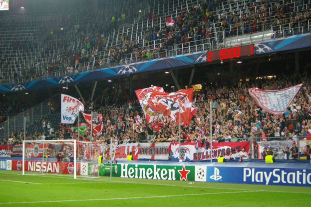 Ein Fußballspiel in einem hell erleuchteten Stadion mit einer großen Zuschauermenge, grünem Rasen, einem Torpfosten mit Bannern und einem Bildschirm in der oberen linken Ecke.