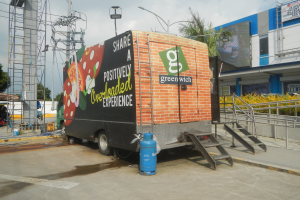 Lastwagen vor einem Gebäude mit einer Tafel, auf der "Teilen Sie eine positiv überladene Erfahrung" steht, mit Stufen, einem Zylinder auf dem Boden, Geländern, Strommasten, Laternen, Bäumen und einem bewölkten Himmel.