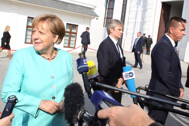 Bundeskanzlerin Angela Merkel spricht mit Reportern ├Ąußerlich der Kanzlei in Berlin, umgeben von Menschen mit Mikrofonen und Geb├Ąuden im Hintergrund.