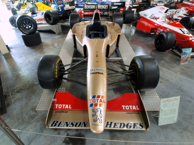 Eine Gruppe von Rennwagen in einem Museum ausgestellt, getrennt durch einen Metallzaun auf der linken Seite, mit einer Texttafel und Menschen im Hintergrund.